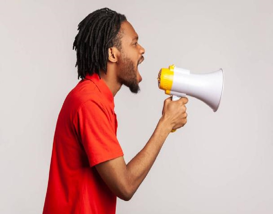 Profile of man with dreadlocks and beard wearing red casual style T-shirt, loudly screaming at megaphone, making protest, wants to be heard. Indoor studio shot isolated on gray background.